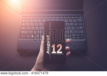 August 12nd. Day 12 Of Month, Calendar Date. Hand Holding Mobile Phone On Laptop Computer Dark Backg