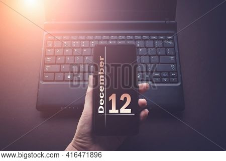December 12nd. Day 12 Of Month, Calendar Date. Hand Holding Mobile Phone On Laptop Computer Dark Bac