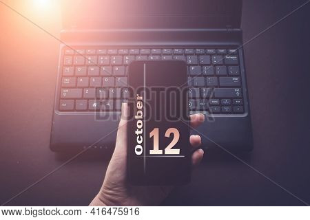 October 12nd. Day 12 Of Month, Calendar Date. Hand Holding Mobile Phone On Laptop Computer Dark Back