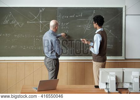 Mature bald professor and young mixed-race student discussing solution of equation by blackboard while teacher pointing at one of its derivative