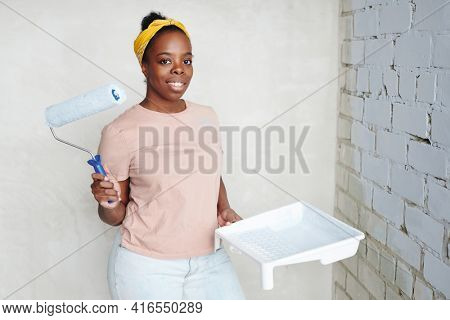 Young smiling woman of African ethnicity with paintroller standing in the corner against two walls while painting it into white color