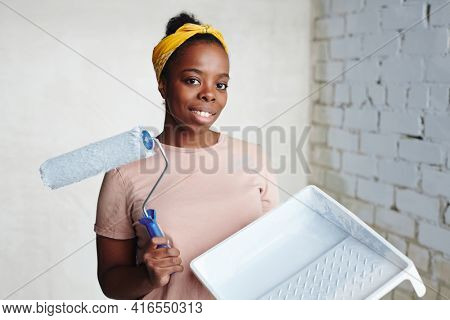 Young cheerful African woman with paintroller standing in the corner against two walls and looking at you with smile during home renovation