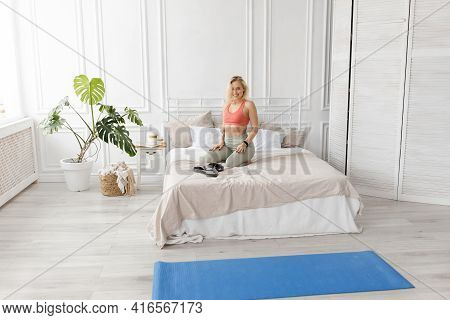 Young Blonde-hair Woman In Sportswear Is Sitting On A Bed, Getting Ready For Morning Workout. Sporti