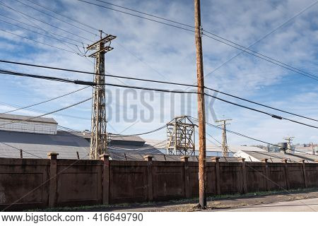 Industrial Steel Power Towers And Warehouses Behind A Tall Concrete Wall, Blue Sky With Clouds, Hori