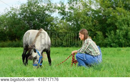 Young Caucasian Woman And Her Hungry Pony In The Field, Side View.