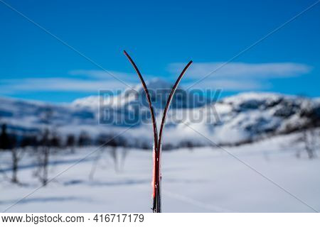 Skis Stacked Together In The Snow With Blurred Snow Capped Mountains.