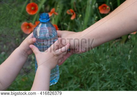 The Child's Hands Take An Open Bottle Of Water From The Right Hand Of The Mother's Hands On A Backgr