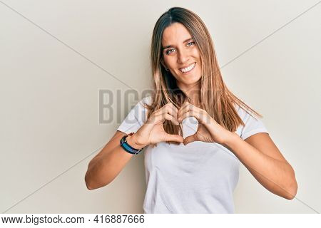 Brunette young woman wearing casual white t shirt smiling in love doing heart symbol shape with hands. romantic concept. 