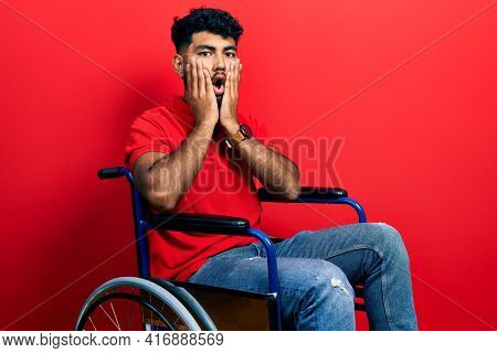 Arab man with beard sitting on wheelchair afraid and shocked, surprise and amazed expression with hands on face 