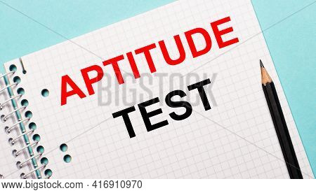 On A Light Blue Background, A Checkered Notebook With The Words Aptitude Test And A Black Pencil.