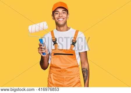 Young handsome african american man wearing cap and painter clothes holding painting roll looking positive and happy standing and smiling with a confident smile showing teeth 