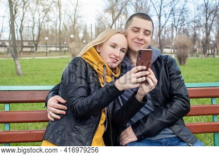 Husband And Wife Take A Selfie While Sitting On A Swing In The Park. Happy Couple Taking Pictures Of