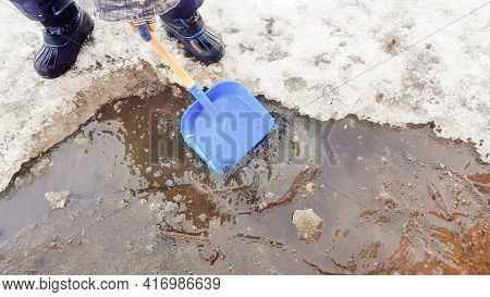 Close-up Of A Child's Feet In Rubber Boots, A Boy Child Happily Playing With A Shovel In A Muddy Pud