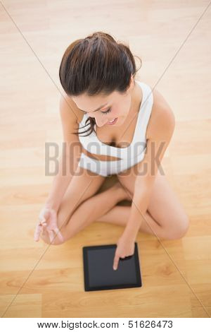 Fit woman sitting in lotus pose using her tablet pc on parquet floor at home