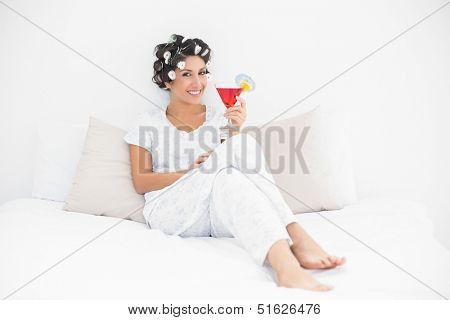 Smiling brunette in hair curlers sitting on her bed holding a cocktail in bedroom at home