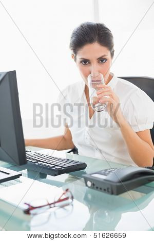 Stern businesswoman drinking a glass of water at her desk in her workplace