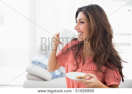 Happy brunette sitting on her sofa holding cup and saucer with a cookie at home in the sitting room