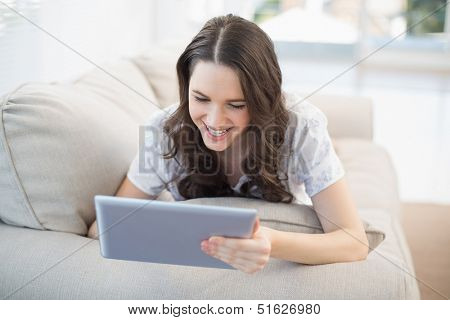 Cheerful woman lying on a cosy couch in bright living room using tablet pc