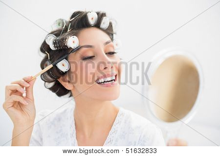 Smiling brunette in hair rollers looking in hand mirror and brushing her eyebrows at home in bedroom