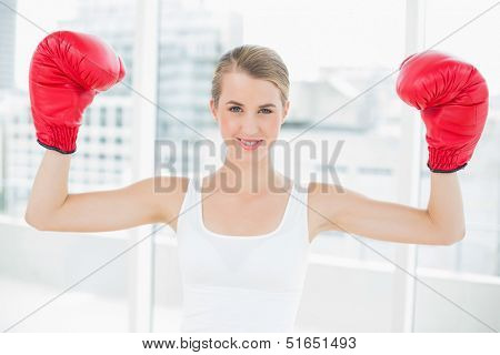 Smiling competitive woman with red boxing gloves cheering up in bright sports hall