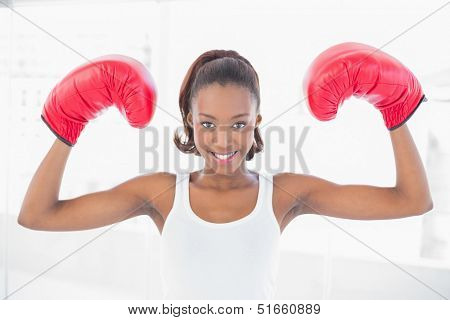 Slender athletic woman wearing boxing gloves making victorious gesture in bright fitness studio