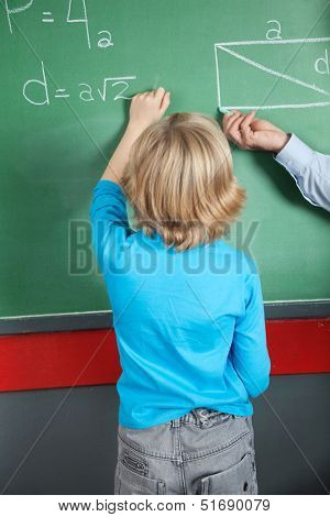 Rear view of little boy writing formula on greenboard in classroom