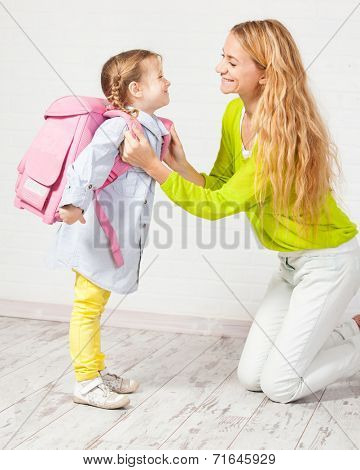 Mother helps her daughter get ready for school. Mom support child to wear a backpack