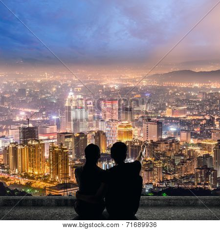 Silhouette of couple sit on ground point faraway on the roof above the city in the night.