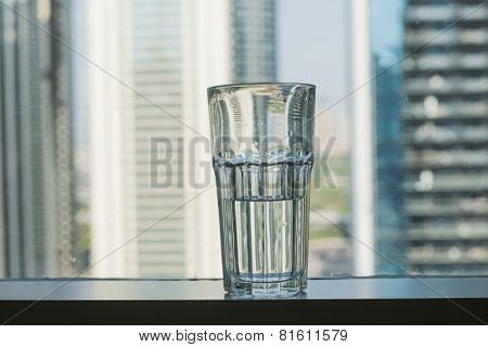 A half filled glass of water placed against window with skyline background. 