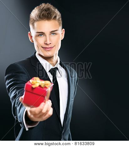Young handsome man giving red gift box. Elegant businessman in dark suit over black background. Sexy blonde smiling guy with holiday Valentine's Day gift