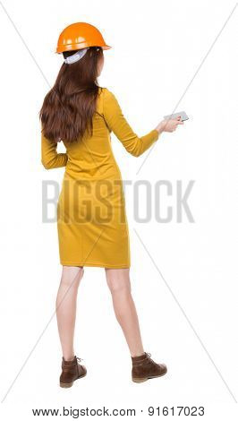 Backview of business woman in construction helmet stands and enjoys tablet or using a mobile phone.  backside view of person. Girl engineer in the yellow dress holds someone plate.