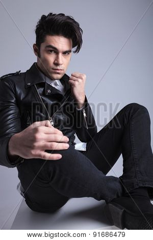 Young fashion man sitting with his legs crossed on grey studio background. He is holdinh one hand on his knee.