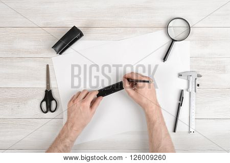 Workplace of draftsman equipped with pencil, ruler, pen, stapler, scissors and magnifying glass. Hands of man holding centimeter ruler and draw on white paper. Top view.