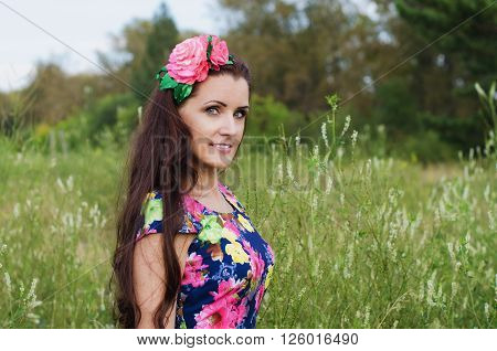 beautiful woman with long hair decorated with artificial flowers stands in a field among the tall grass on a summer evening