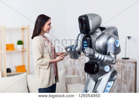 Help me. Cheerful positive charming girl using tablet  and smiling  while giving it to the robot