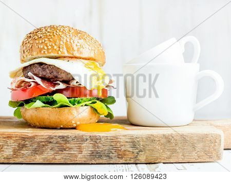 Breakfast set. Homemade beef burger with fried egg and vegetables, onion rings and coffee cups on wooden board, white painted background. Selective focus