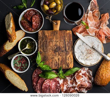 Wine snack set with empty wooden board in center. Glass of red, meat selection, mediterranean olives, sun-dried tomatoes, baguette slices, camembert cheese and spices on black background, top view, copy space