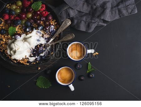 Healthy breakfast. Oat granola crumble with fresh berries, seeds and ice-cream in iron skillet pan, coffee cups on dark wooden board over black backdrop, top view, copy space