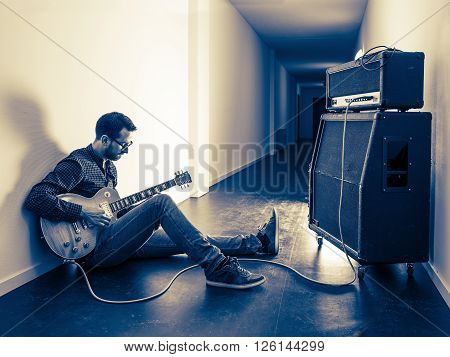 Photo of a man sitting playing his electric guitar in front of a large amplifier in a hallway. Filtered for retro vintage feel.