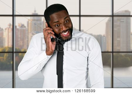 Excited darkskinned guy holding cellphone. Friendly executive near office window. His team is making progress. You can count on me.