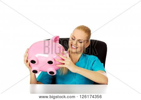 Young female doctor or nurse sitting behind the desk and holding piggybank