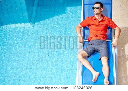 young man relaxing on the chaise-longue near the pool. man wearing in red polo shirt and sunglass