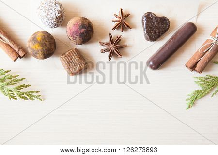 Cuisine background. Six raw candies made of date fruit nuts hibiscus curry cocoa coconut decorated with cinnamon sticks cardamom thuja branches on light wooden background indoors. Healthy and raw sweet-shop design content.
