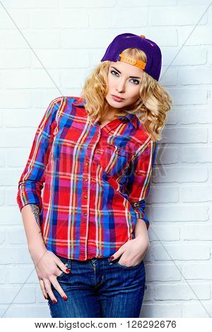 Beautiful modern girl with curly blonde hair wearing baseball cap and checkered shirt posing by a brick wall. 