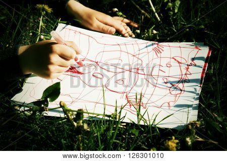 Close Up View Of Child's Hand Drawing Colorful Ornaments On Paper. Notebook In Fresh Green Grass Of