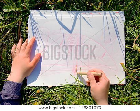 Close Up View Of Child's Hand Drawing Colorful Ornaments On Paper. Notebook In Fresh Green Grass Of