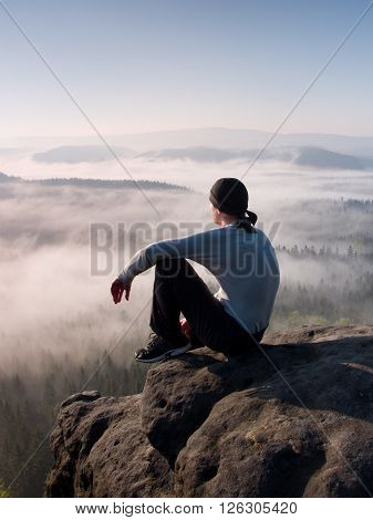 Man sit on the peak of rock and watching into colorful mist and fog in forest valley.