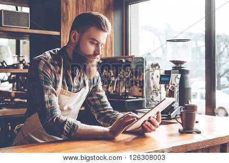Technologies make business easier. Young bearded man in apron using his digital tablet while leaning on bar counter in coffee cafe.