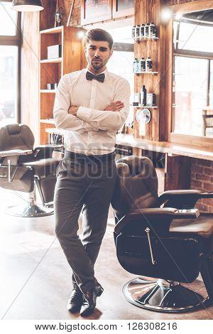 Barber expert. Full length of young handsome man looking at camera and keeping arms crossed while standing at barbershop