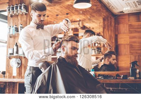 Everything should be perfect. Side view of young bearded man getting haircut by hairdresser while sitting in chair at barbershop
